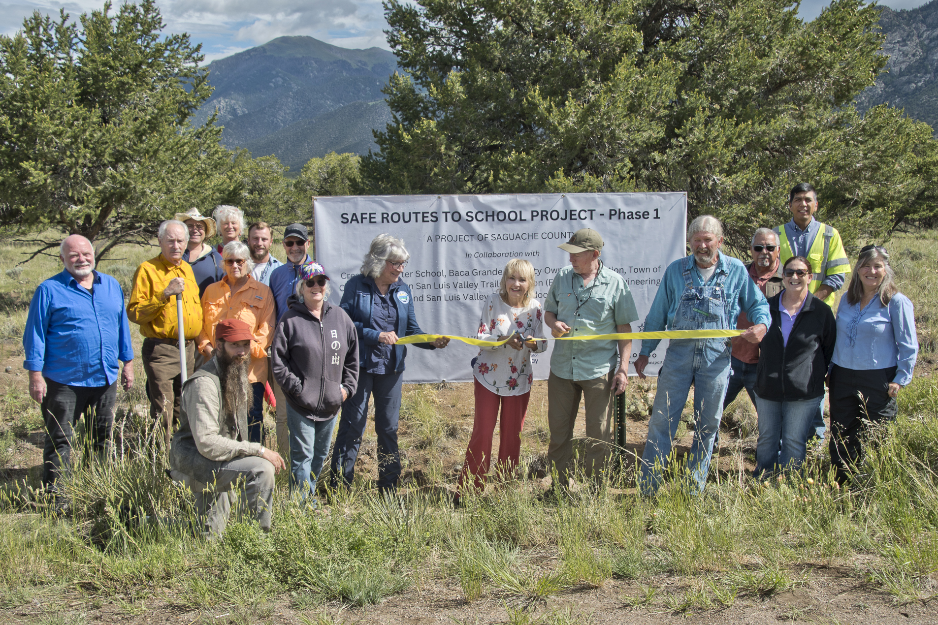 A group of individuals stands outdoors holding a banner that reads “SAFE ROUTES TO SCHOOL PROJECT – Phase 1” with additional text below that is partially obscured. The group appears to be participating in a ceremonial ribbon-cutting event, The setting includes trees and mountains in the background, suggesting a rural or natural location.