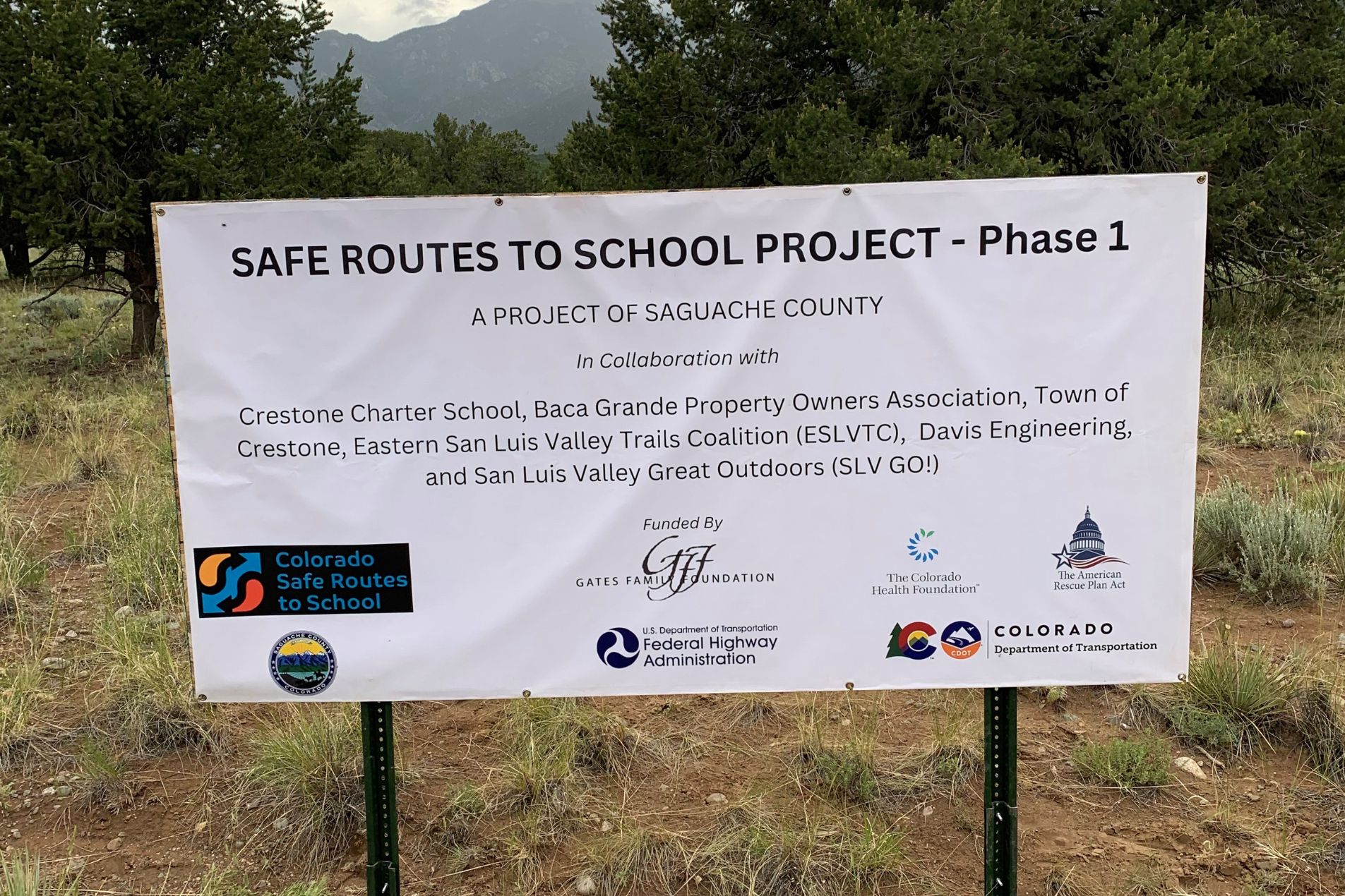 The sign is white with black and green text, and it’s mounted on two metal posts. The backdrop features a natural setting with trees and overcast skies. The sign lists the project collaborators, which include Crestone Charter School, Baca Grande Property Owners Association, Town of Crestone, Eastern San Luis Valley Trails Coalition (ESLVTC), Saguache County Government, and San Luis Valley Great Outdoors (SLV GO!). Each collaborator’s name is accompanied by a logo. 